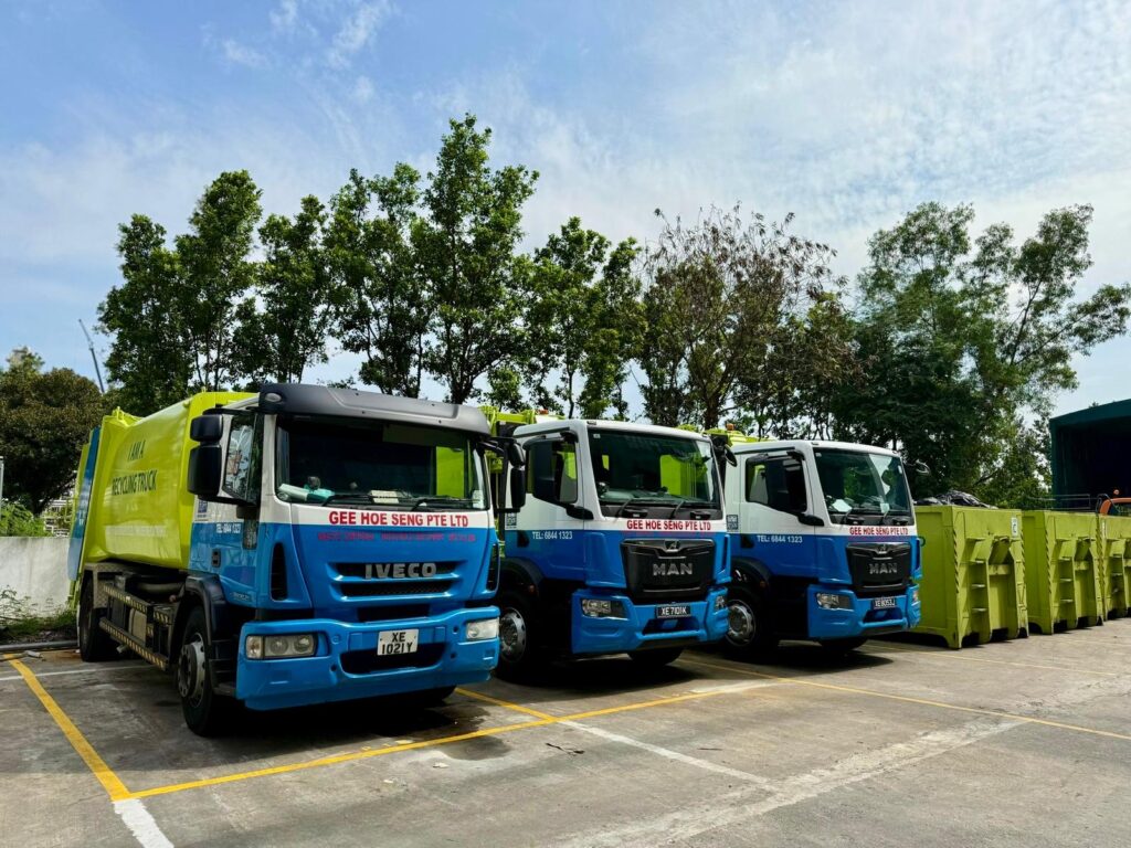 Gee Hoe Seng Pte Ltd recycling and collection trucks parked beside green open top containers at operations facility in Singapore.